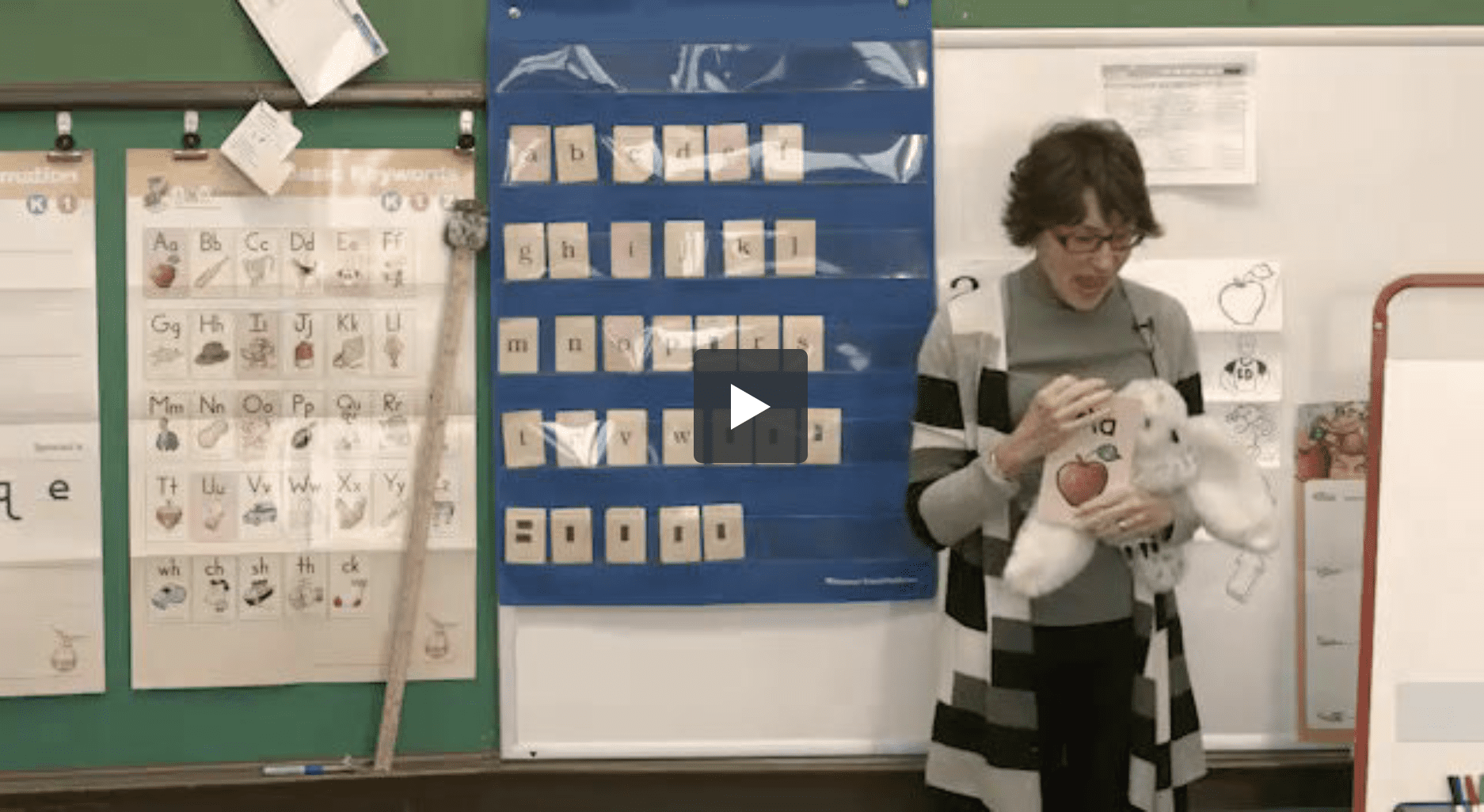 A teacher stands in a classroom holding a stuffed animal. Behind her, alphabet charts, a pocket chart with letter cards, and a green board are visible. The teacher is wearing a grey cardigan and glasses.